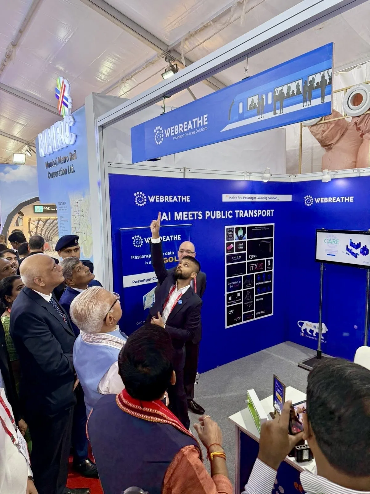 Presenter showing an overhead passenger-counting sensor beside Mumbai Metro Rail signage